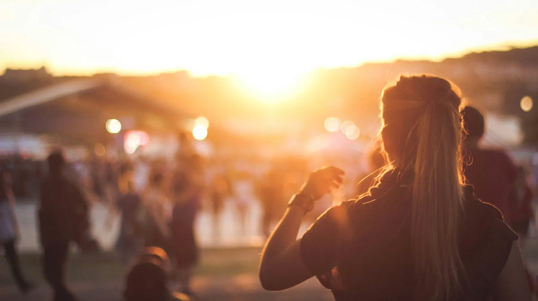 A woman seen from behind in a crowd at an outdoor gathering, facing the setting sun.